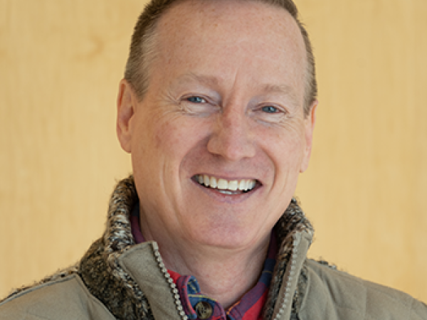 head and shoulders of smiling Troyd Geist wearing tan vest over red flannel shirt, with short brown hair and a cream-colored background