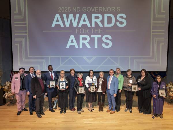 Large group photo with ND Governor's Awards for the Arts on screen above a stage with all seven recipients and ND Governor and First Lady Armstrong