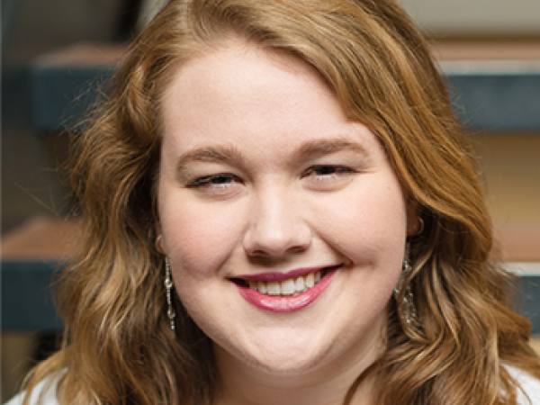 head and shoulders of Brenna Lahren with long, curly, red-brown hair, light skin, toothy smile with red lipstick, long dangling earrings, light grey shirt with blurred stairwell in the background