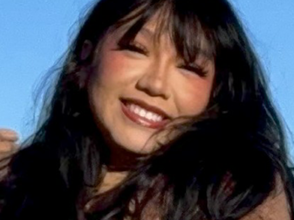 head and shoulders of Kiera Fox with long, straight, black hair, toothy smile with dark red lipstick, light brown jacket with dark brown under shirt and a blue sky and canyon background