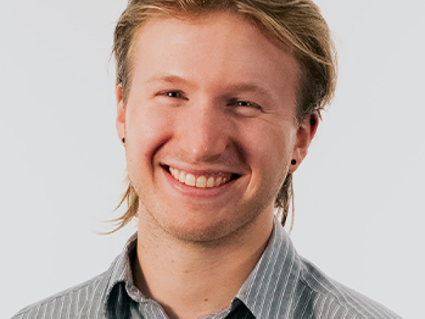head and shoulders of Tobias Zikmund with short, dirty blonde hair, black dot earrings, pale skin and a big toothy smile; wearing a grey collared shirt with white stripes