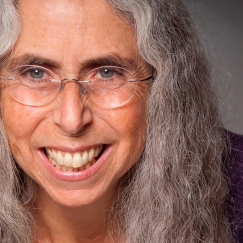 Close-up headshot of Beth Olshansky with big smile, clear glassses and long gray curly hair