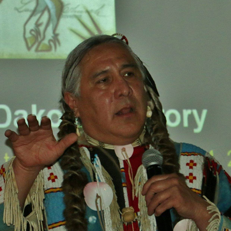 Head and shoulders of Monte Yellowbird in native apparel, talking on microphone w/ native horse image on slide projected behind him