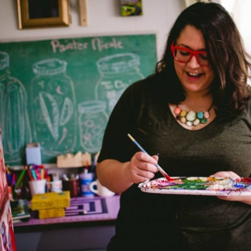 Teaching artist Nicole Gagner mixing paint, wearing red-rimmed glasses, with long, thick wavy dark hair, enjoying herself
