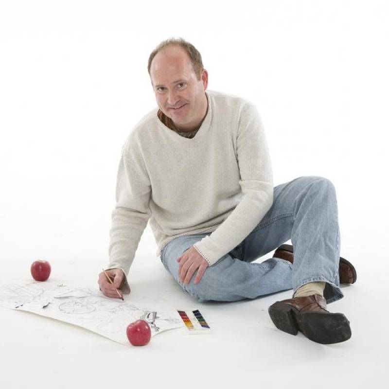 Teaching artist Markus Tracy with balding brown hair, smiling brightly, sitting on a white floor with crossed legs, wearing blue jeans and a white sweater, playing a game with red balls