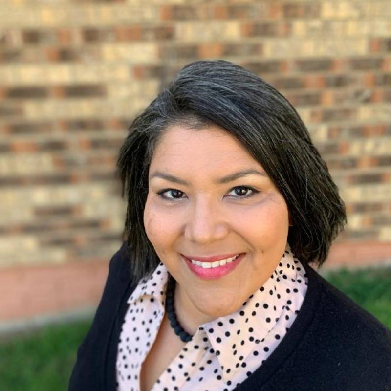head and shoulders of teaching artist Shelly Sayler with short, straight black hair, wearing a black sweater with a white patterned undershirt, black necklace and toothy smile
