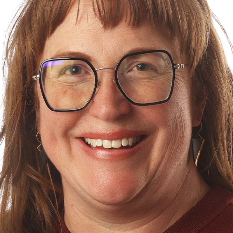 Ana Paige with red sholder length straight hair smiles into the camera with large black glasses and gold triangle earrings