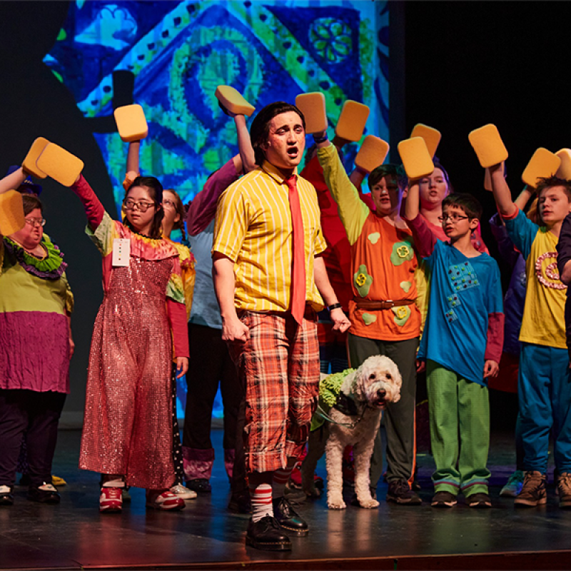 Colorful cast of various races, ages, and abilities in The SpongeBob Musical inhabit the stage in all their glory with neurodiverse SpongeBob and Deaf Patrick Star gesturing grandly with arms open. Photo Credit: Jeff Heimbach