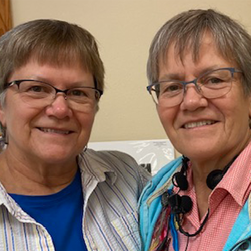 Head and shoulders of sisters Barbara and Beverly Benda, with short brown/grey hair, matching cat-style glasses and toothy smiles