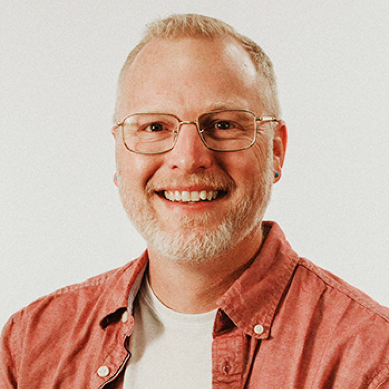 Head and shoulders of Matthew Anderson wearing a dark coral button-up shirt with a white undershirt, clear rimmed glasses, with pale skin, a big toothy smile, dark eyes, short, light hair and short gray beard