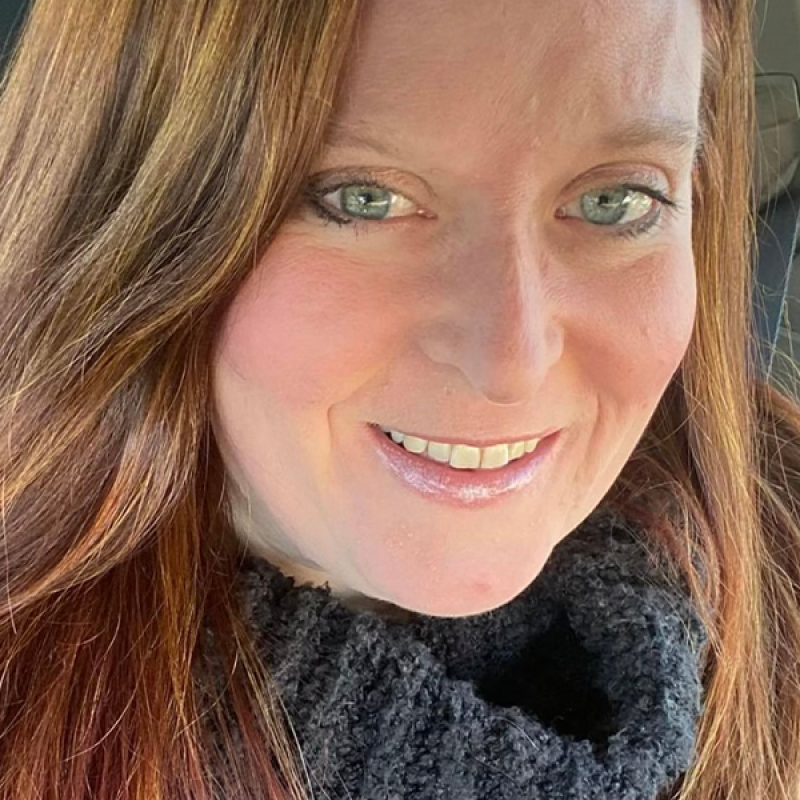 close up face and shoulders of artist Myra Klein with long, straight auburn hair, blue eyes, heavy grey turtle-neck sweater, pale skin, and slightly toothy smile
