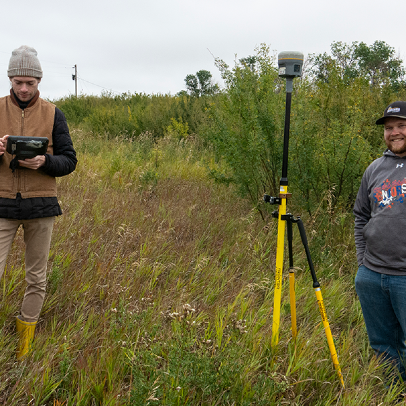 Two men standing outside on an overgrown field on a fall day with surveying equipment