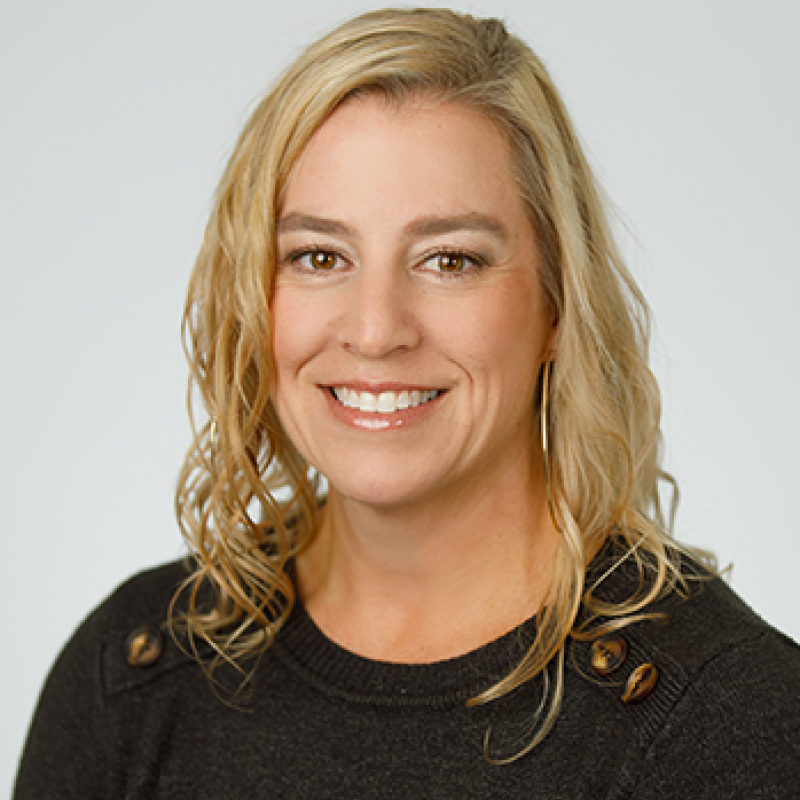 Head and shoulders of Rhea Beto with brown eyes, curly, shoulder-length blond hair, light skin, toothy smile, wearing dark long-sleeve shirt with light grey background