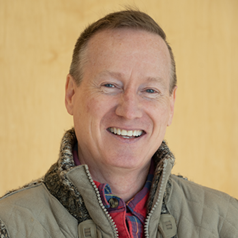 head and shoulders of smiling Troyd Geist wearing tan vest over red flannel shirt, with short brown hair and a cream-colored background