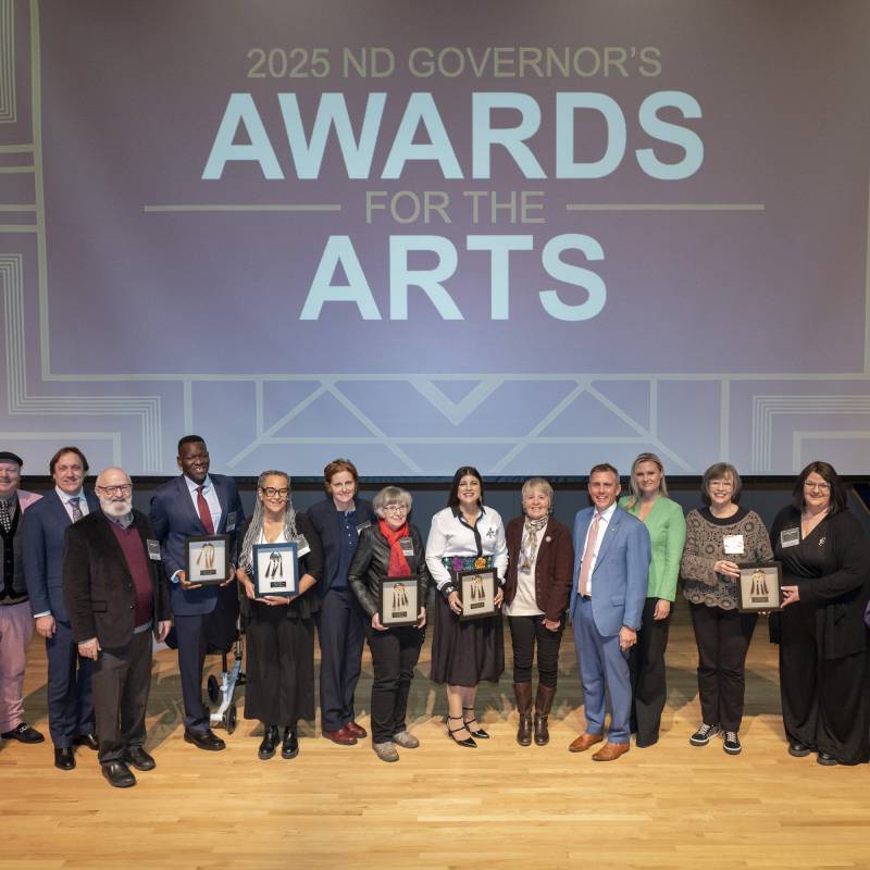 Large group photo with ND Governor's Awards for the Arts on screen above a stage with all seven recipients and ND Governor and First Lady Armstrong