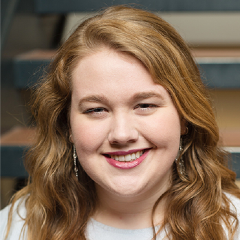 head and shoulders of Brenna Lahren with long, curly, red-brown hair, light skin, toothy smile with red lipstick, long dangling earrings, light grey shirt with blurred stairwell in the background