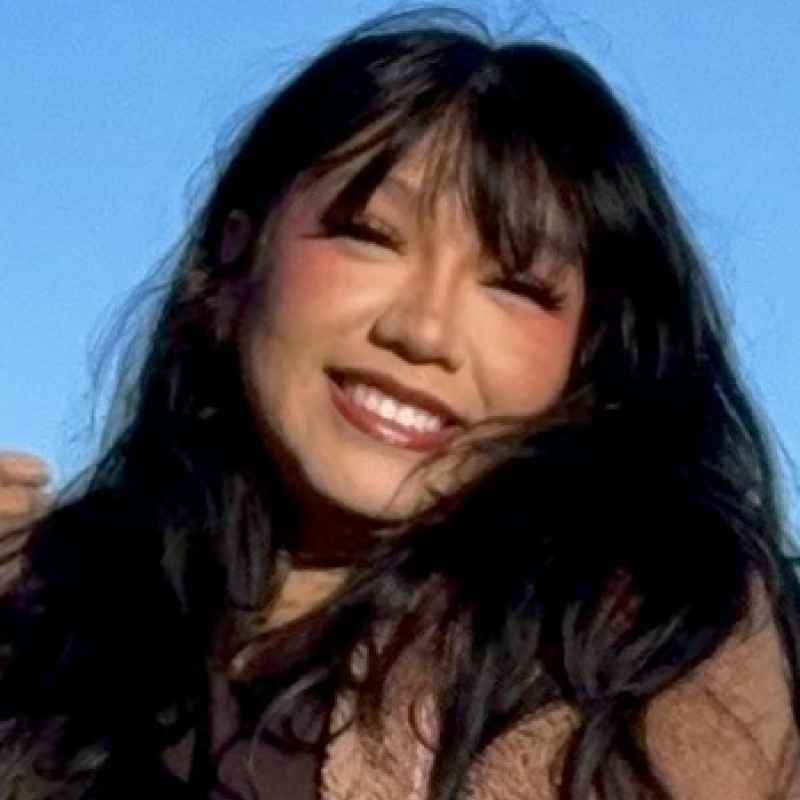 head and shoulders of Kiera Fox with long, straight, black hair, toothy smile with dark red lipstick, light brown jacket with dark brown under shirt and a blue sky and canyon background