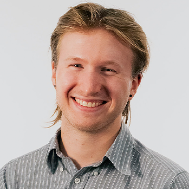 head and shoulders of Tobias Zikmund with short, dirty blonde hair, black dot earrings, pale skin and a big toothy smile; wearing a grey collared shirt with white stripes