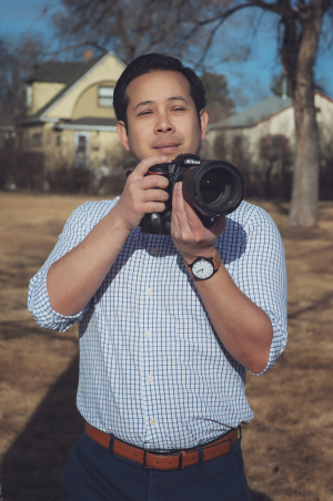 Waste up photo of John Geyerman, Asian with short dark hair, jeans and white button-down shirt with light blue grid lines, holding large camera, standing in big open field early spring