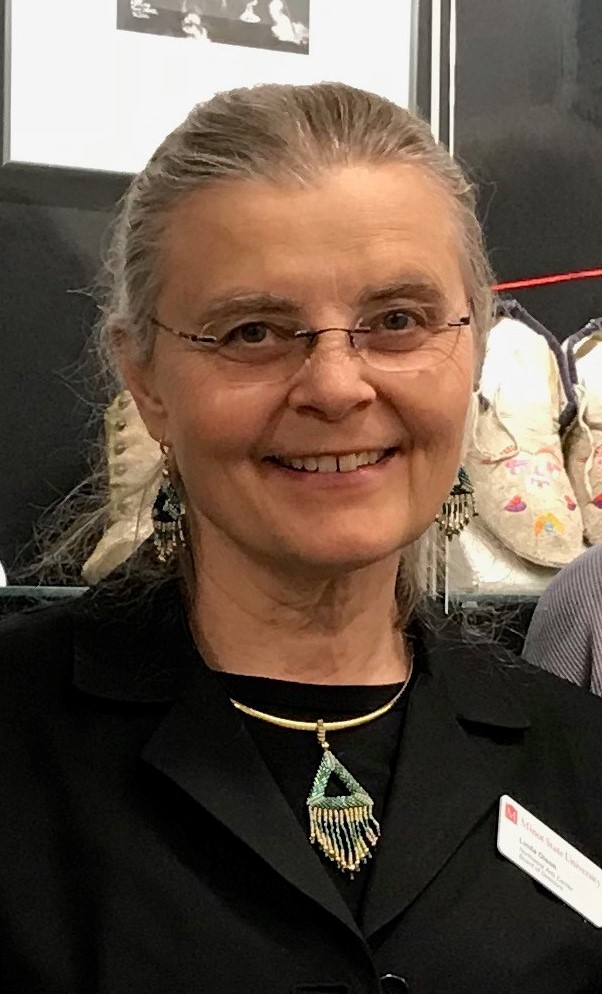 Linda Olson with dirty grey hair pulled back into a ponytail with clear rimmed glasses and a black suit jacket