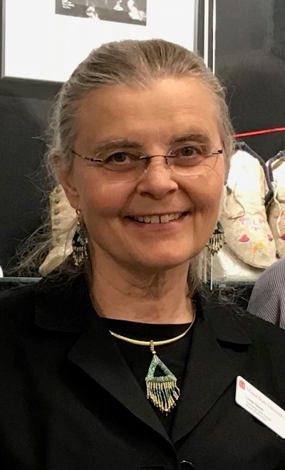 Head and shoulders of Linda Olson with dirty grey hair pulled back into a ponytail with clear rimmed glasses and a black suit jacket