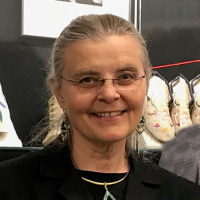 Head and shoulders of Linda Olson with dirty grey hair pulled back into a ponytail with clear rimmed glasses and a black suit jacket