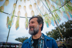 Head and shoulders of James Peterson with thick salt and pepper hair and beard, wearing a jean jacket with a large public outdoor artwork behind him