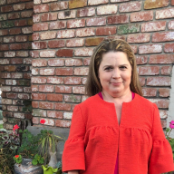 Waste-up photo of Katharine Hayward with apple red shirt, long, straight brown hair, pale skin, standing outside in front of brick wall