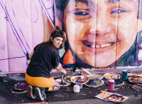 Greta McLain kneeling in front of huge outdoor mural she is painting with mess of paints and brushes all around her