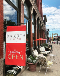 Dakota Fine Art Gallery Open sign in front of a brick building with many windows and comfy chairs in front on a summer day
