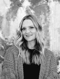 Greyscale, waste-up image of artist Nancy Ness wearing a light sweater cardigan with a black shirt underneath, standing in front of her artwork, with long, wavy blonde hair and a slightly toothy smile