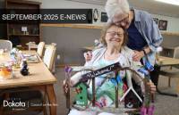 An older woman with white hair embraces another woman with glasses who is smiling and holding a decorative wooden sign. The sign features birdhouses and butterflies, with text that reads "Welcome," "Bless," "This," and "Nest." In the background, there are tables with crafting supplies and various finished craft items. The image also includes text overlays: "SEPTEMBER 2025 E-NEWS" at the top and "NORTH Dakota | Council on the Arts Be Legendary." at the bottom left. 