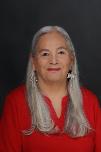 Dr. Denise Lajiimodiere, closed smile, wearing a bright red shirt, dangling dragonfly earrings with long grey hair