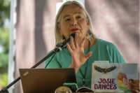 Dr. Denise Lajiimodiere, with long grey hair, large earring and hanging necklace, standing at a podium with a microphone, reciting from her book Josie Dances