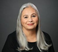 Head and shoulders of Dr. Denise Lajimodiere with long, straight, grey hair and a closed smile; wearing a black scoop-neck shirt