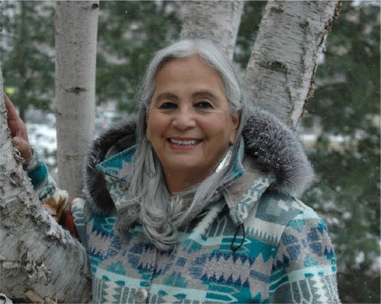 Dr. Denise Lajiimodiere, big toothy smile, long grey hair, wearing a bright-colored Tribal coat outside during a cold winter day, standing at the base of large birch trees
