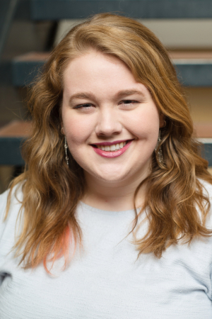 head and shoulders of Brenna Lahren with long, curly, red-brown hair, light skin, toothy smile with red lipstick, long dangling earrings, light grey shirt with blurred stairwell in the background