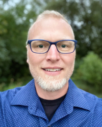 Head and shoulders of Matthew Anderson with royal blue shirt, short blonde hair and beard, blue-rimmed glasses, blue eyes, slight toothy smile and blurred out lush green trees in the background