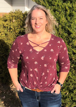 waist-up photo of Rhea Beto wearing burgundy 3/4 length sleeve shirt with flower pattern and criss-cross strings on the chest; she has curly, shoulder-legth dirty blond hair, brown eyes and a big toothy smile; dark green bushes in the background