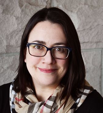 Head and shoulders of Robin Bosch with dark-rimmed glasses, shoulder-length straight dark hair, dark eyes and a closed smile, wearing a dark shirt with a plaid patterned scarf on a textured off-white background