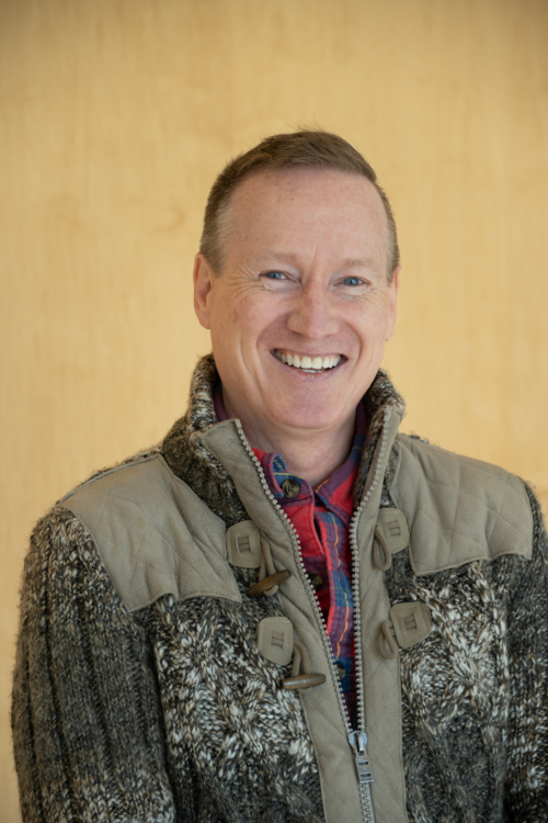 head and shoulders of Troyd Geist wearing a tan vest over a red flannel shirt with short brown hair, blue eyes, a big toothy smile on a cream-colored background