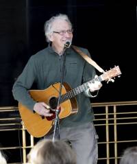 knee-up photo of grey-haired, pale-skinned, Chuck Suchy, smiling while playing an acoustic guitar and singing into a microphone, wearing a green-grey button-up shirt and grey cargo pants