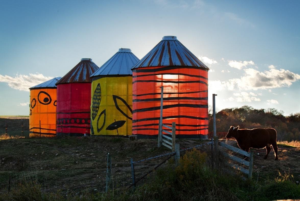 A photo of 4 small brightly colored grain silos standing on a dark brown pasture in Sauk County, rural Wisconsin painted by E. Baillies with corn cribs painted by Brenda Baker.