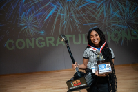 Gabrielle Johnson of Minot High School holding a large trophy, standing in front of a wall that reads CONGRATULATIONS with fireworks exploding all around