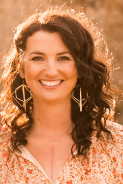 Head and shoulders of Jessie Veeder with sun shining on the back of her flowing, long, dark, curly brown hair, paisley orange flowers on white shirt, big toothy smile, dark brown eyes, big dangly earring in the shapes of diamonds and hexagons, and a visible scar running down her chest where she had surgery.