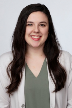 Waste-up photo of Maddi Menich with off-white suit jacket, forest green undershirt, long dark brown wavy hair, toothy smile, pale skin on plain white background