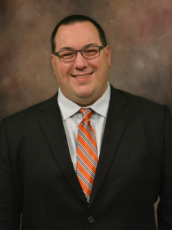 Waste up photo of Russell Mills wearing black suit jacket with orange and grey striped tie and white undershirt. He has a dark black, military cut with skinny black-rimmed glasses, slight toothy grin on brown-toned portrait-style background.