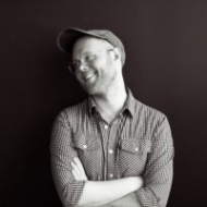 Waste up, greyscale photo of Scott Arley looking left, folded arms, toothy smile, clear glasses, baseball cap, striped, button-up, long-sleeve shirt
