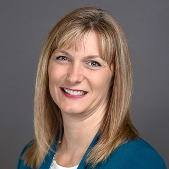 Head and shoulders of straight, blonde-haired, brown-eyed Christi Stonecipher on grey background wearing teal jacket top