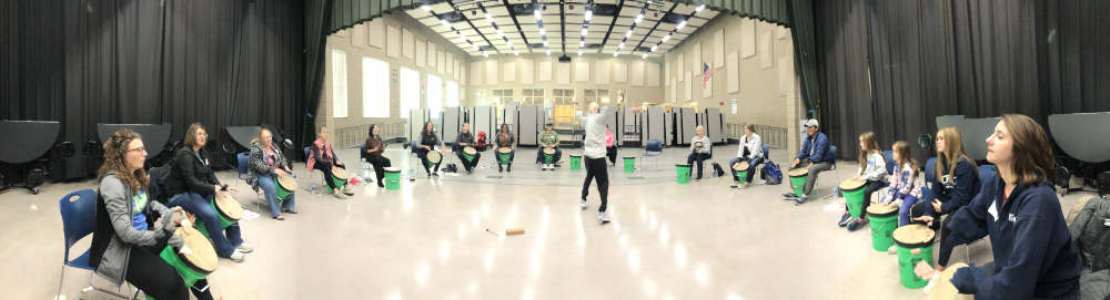 Beat the Odds drumming workshop showing a panoramic about 17 people drumming on 5 gallon buckets with lids while a person in the center directs them